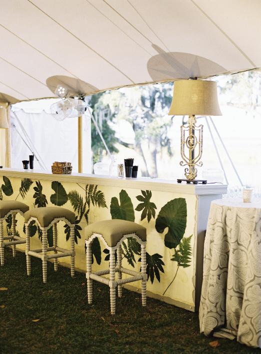For a stunning and singular bar setup in keeping with the wedding’s aesthetic, planner Calder Clark affixed fern, fatsia, and elephant ear leaves to linen fabric, then pressed them behind a glass front. Custom bar by Blossom Events. Bar stools and lamps by Calder Clark. Linens by La Tavola. Tent by Sperry Tents Southeast. Photograph by Tec Petaja.