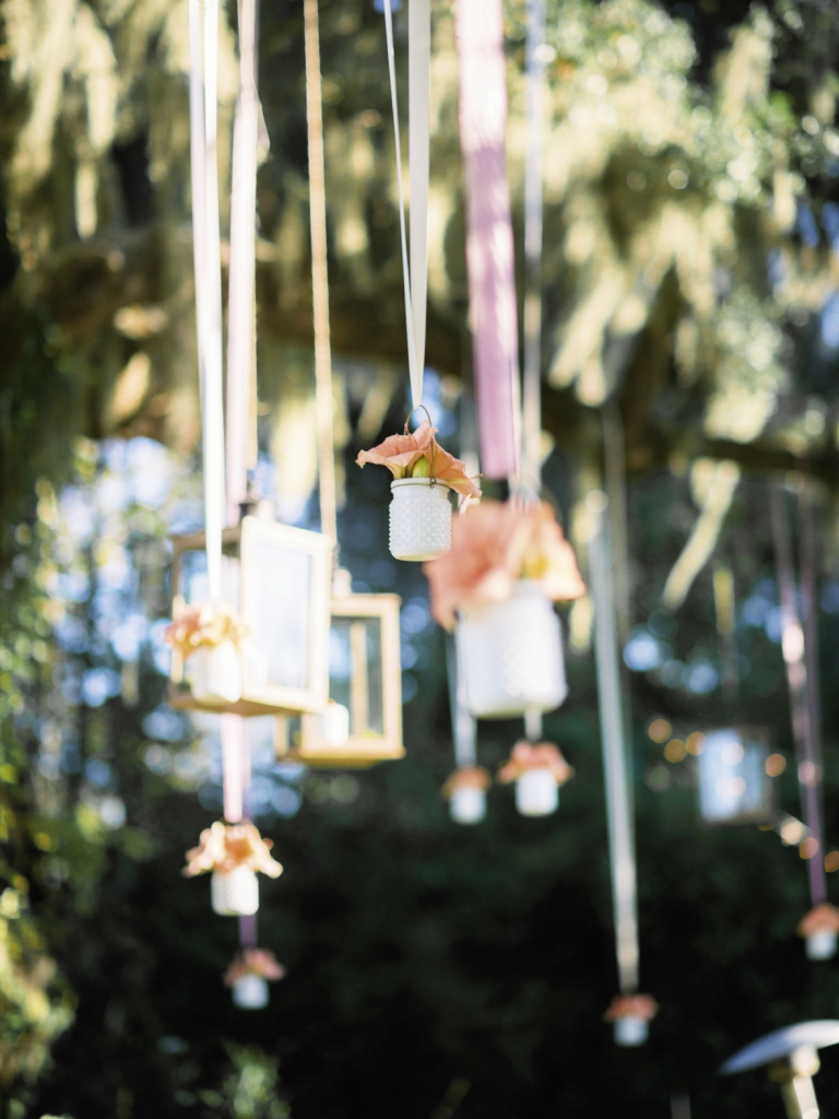 FAIRY TALE: To frame the couple’s big moment, Whitney Randall of Branch Design Studio tucked coral amaryllis blooms into opaque milk glass jars and hung them among glass lanterns in the live oak above the altar.