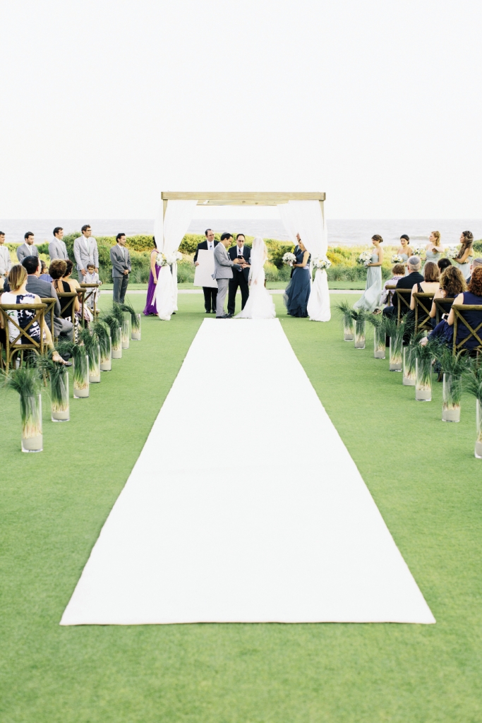 WILD IDEAS: The Sanctuary Hotel’s immaculate lawn made the pearl-white aisle runner a pristine  possibility. Bear grass bedded in sand-filled glass  cylinders lined the aisle. The rustling blades danced in the wind and harkened the nearby dune vegetation.